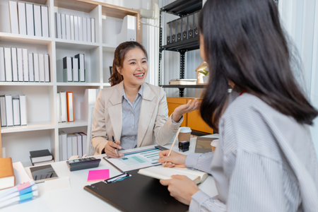 Two Asian businesswomen discussing ideas and analyzing reports from documents in a modern office. They collaborate on business strategies, teamwork, and organizational planning for success.の写真素材