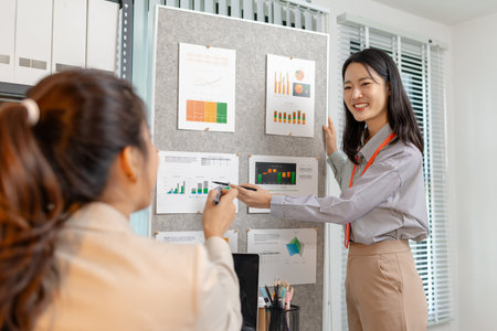 Two Asian businesswomen are smiling while discussing company performance reports in a modern office....