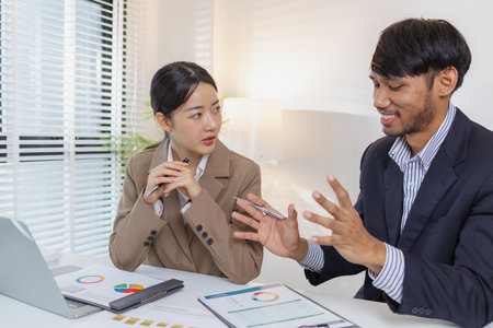 Two business professionals are discussing financial charts and reports in a modern office. They collaborate, share ideas, and analyze data together, highlighting teamwork and strategy.の写真素材