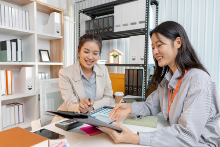 Two Asian businesswomen discussing ideas and analyzing reports from documents in a modern office. They collaborate on business strategies, teamwork, and organizational planning for success.の写真素材