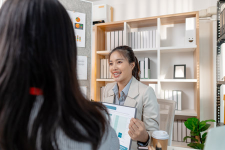Two Asian businesswomen discussing ideas and analyzing reports from documents in a modern office. They collaborate on business strategies, teamwork, and organizational planning for success.の写真素材