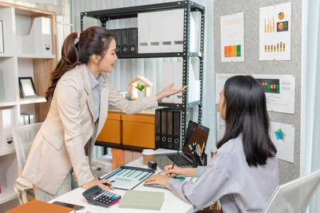Two Asian businesswomen are smiling while discussing company performance reports in a modern office. They analyze data from printed documents, showing teamwork, planning, and collaboration in businessの写真素材