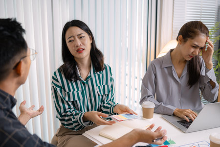 A tense business meeting where a team discusses design problems and project challenges. The man tries to explain solutions while one team member looks stressed, showing workplace pressure and teamworkの写真素材