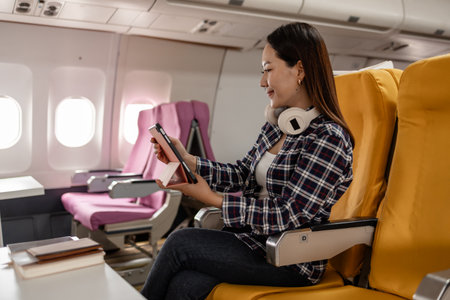 Young Asian woman sitting in airplane seat with yellow cover, smiling while using tablet during flight. She wears casual outfit with headphones around her neck, enjoying digital entertainment.の写真素材