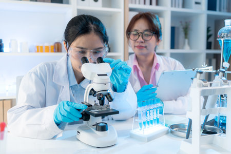 Two female scientists in white lab coats working together in a bright laboratory with microscopes,...