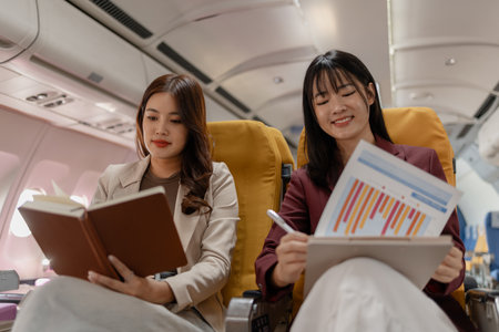 Two women sit side by side on an airplane, reviewing documents together with focused smiles,...