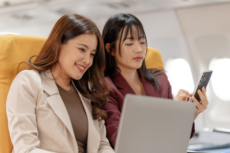 Two women sit side by side on an airplane, reviewing documents together with focused smiles, representing teamwork, business discussion and productive collaboration during calm and comfortable flight.の写真素材