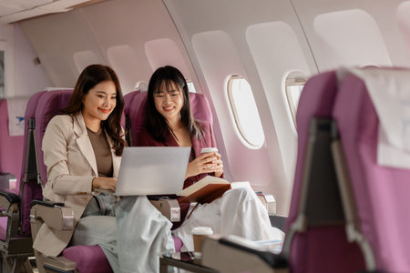 Two women sit side by side on an airplane, reviewing documents and working on a laptop together,...