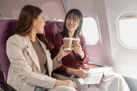 Two women sit together on an airplane, smiling and holding cups of coffee by the window, expressing...