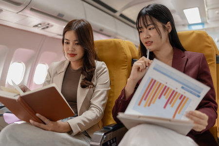 Two women sit side by side on an airplane, reviewing documents together with focused smiles, representing teamwork, business discussion and productive collaboration during calm and comfortable flight.の写真素材