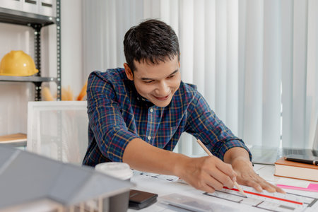 A young male architect is working with blueprints at his desk, using rulers and drawing tools to...