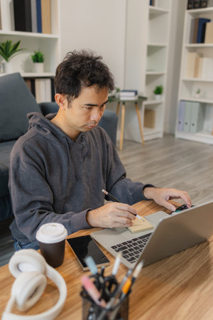 Man calculating expenses while working on laptop at home desk with calculator and smartphone,...