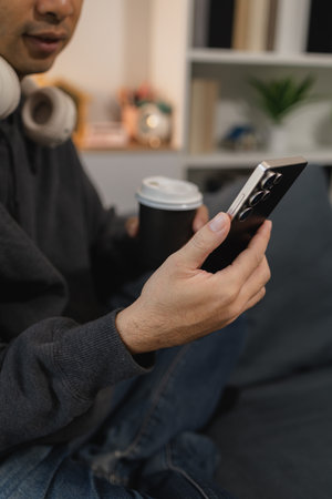 Man using smartphone while relaxing on sofa at home, browsing mobile apps and digital content,...