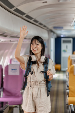 Young female traveler boarding an airplane, placing carry on luggage in overhead compartment, checking smartphone and passport while finding her seat before departure for her journey.