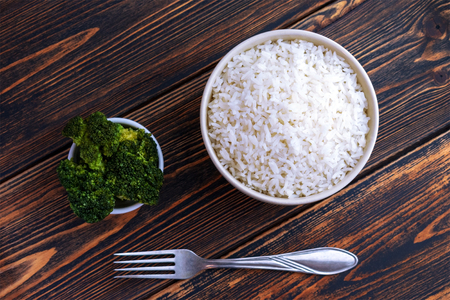 A Cup of white Rice and broccoli on a dark wooden background. Top view. Dietary or vegetarian food. Healthy and proper nutrition. Carbohydrates used by athletes in preparation for the competition. Food cooked at home. On the table lies a fork.の写真素材