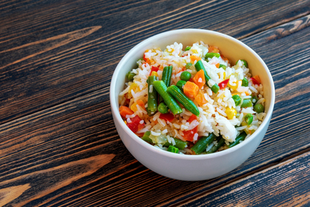 A Cup of white rice, green beans, corn, pepper, peas, carrots and onions on a dark wooden background. Dietary or vegetarian food. Healthy and proper nutrition. Food cooked at home. Location on the right side.の写真素材