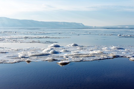 Beautiful winter or spring landscape. Breaking ice on the river. In the background in the distance you can see the mountains covered with fog. Sunlight breaks through the clouds in the blue sky and reflects off the water.の写真素材