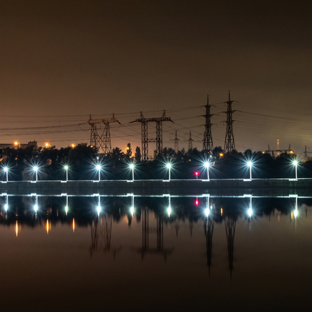 Electric towers are beautifully reflected in the water at night. Concrete dam on which the street lights are arranged in a row. Behind the dam trees and electric high-voltage towers of different typesの写真素材