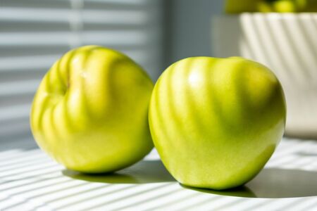 Two green apples on the table next to the window. In the background, a bowl of apples. Illuminated by sunlight passing through the closed blinds. close-up photo. photo in light colors.の写真素材