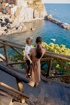 A beautiful woman holding her little child in her arms, admiring the colorful village of Manarola in 5 Terre. The stunning scenery of the Mediterranean coast, the old town, the port, the terraces, and the vineyards, create a perfect setting for a relaxing and romantic vacation.の写真素材
