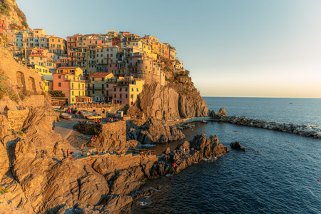a breathtaking sunset view of the charming village of Manarola in Italy's 5 Terre region. The warm golden hues of the sun perfectly complement the colorful buildings, creating a stunning contrast against the deep blue waters of the Mediterranean Sea.の写真素材
