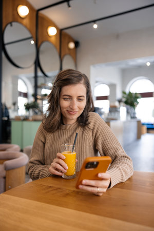 Portrait of a smiling woman using mobile phone while drinking orange juice in cafeの写真素材