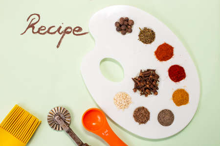 Multiple spices (paprika, curcuma, all spice, cloves, pepper) on a white painter's palette, with Recipe word written with brown chalk on light green paperの写真素材