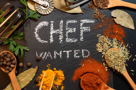 Chef wanted written on black stone tile with white chalk, surrounded by condiments an kitchen tools (paprika, curcuma, all spice, parsley, wood spoonsの写真素材