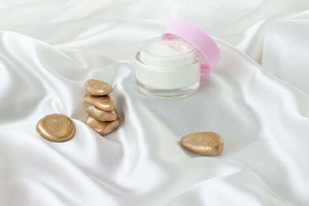 Glass jar with visible white cream and opened pink lid next to a stack of golden stonesの写真素材