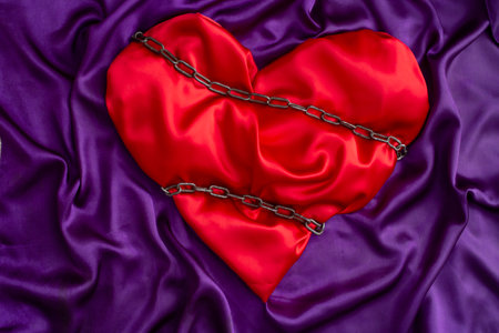 red satin heart tied with chains on purple background, marriage restrictions or jealousy metaphorの写真素材