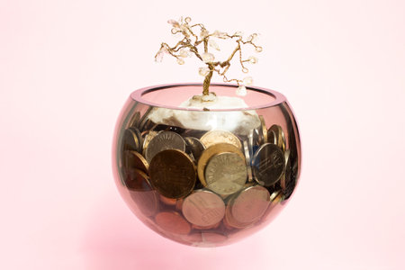 Glass bowl full of coins with a wire tree on top, levitating on pink background.