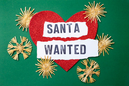Santa wanted written on white paper strips, on a red glitter heart and green background. Santa for...