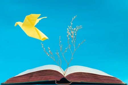 Yellow origami dove flying from an old book on blue background. Soft focus close up of a dry tree growing from a bookの写真素材