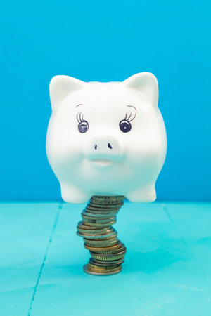 White ceramic piggy bank on a stack of coins, on blue background, soft focus close upの写真素材