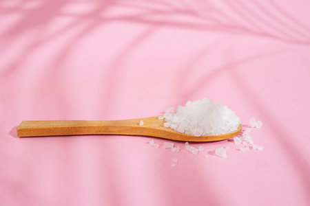 Wooden tea spoon filled with white rock salt, on pink background with palm leaf shadows, soft focus close upの写真素材