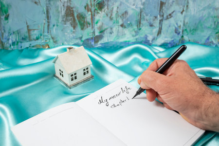 Male hand writing My new life chapter one on a blank notebook diary next to a miniature house model,...