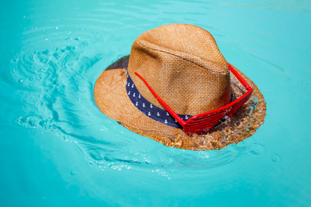 Straw hat with red grated shades splashing in turquoise water surface, abstract