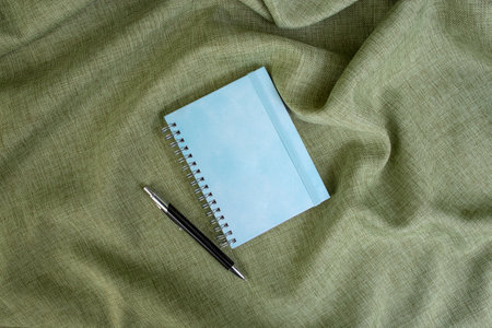 Empty blue hard cover notebook with a ball point pen on olive green fabric backdrop, copy space for textの写真素材
