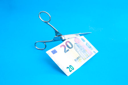 Stainless steel scissors cutting a 20 euro banknote in half on blue background, cutting expenses, crisis metaphorの写真素材