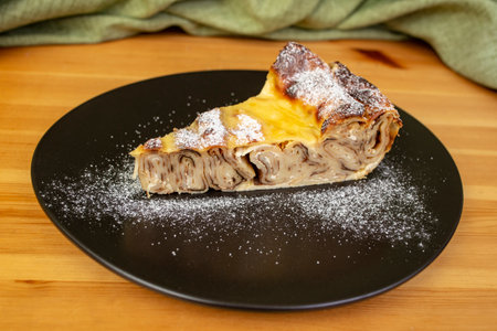 Oven baked rolled pancakes pie slice sprinkle with powder sugar, on a black plate on wooden tableの写真素材
