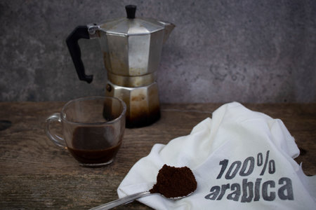 100% arabica written with stamp letters on a white fabric sack next to a mocha cup and a spoon full of ground coffee, soft focusの写真素材