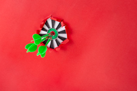 Three green darts in the middle of a target board, thru a ripped hole on red paper background, with copy space for text or productの写真素材