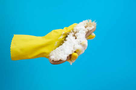 Hand with yellow latex glove holding a hard brush with white foam on blue