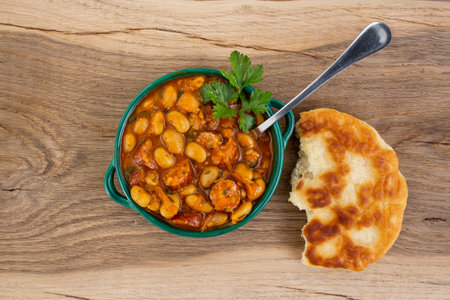 Butter beans stew with tomato sauce and sliced pork sausages with half of a pita bread, on wooden oak board tableの写真素材