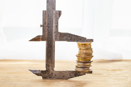 Metal caliper gripping a high stack of golden coins on a wooden desk