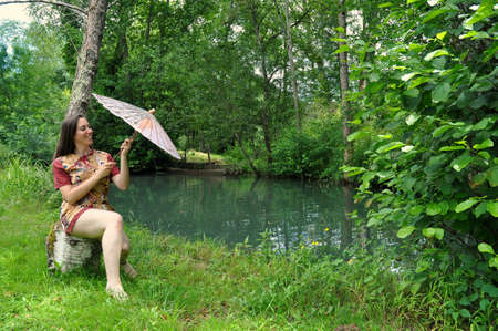 Beautiful young woman with a parasol, sitting in the wilderness waterfrontの写真素材
