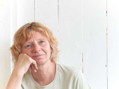 A mature woman leaning against a white fence with a smile, resting cheek on hand.の写真素材