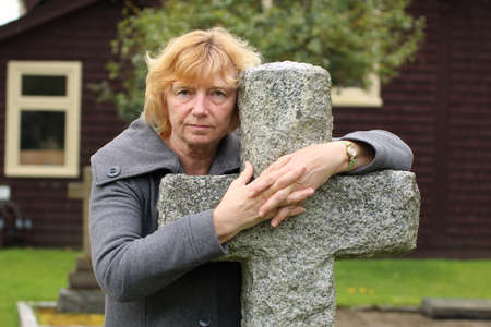 Mature woman praying with hands clasped over a stone cemetery cross, with church in background.の写真素材
