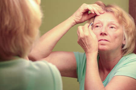Mature woman putting on makeup while looking at mirror reflection.の写真素材
