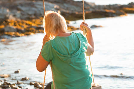 A mature woman sitting on a swing at the beach, her head resting against a swing rope.の写真素材